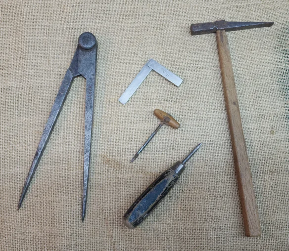 Four vintage hand tools laid on hessian: a compass, try square, awl, and cross-peen hammer