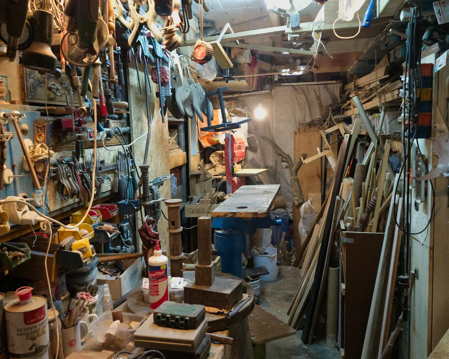 Narrow workshop packed with hanging tools, a drill press at centre, and stacked timber along the right wall